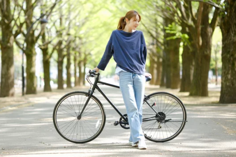新緑の並木道で自転車を引きながら微笑む日本人女性 ─ Mani Padme40代・なりわいと自分のペースで歩む人生の道