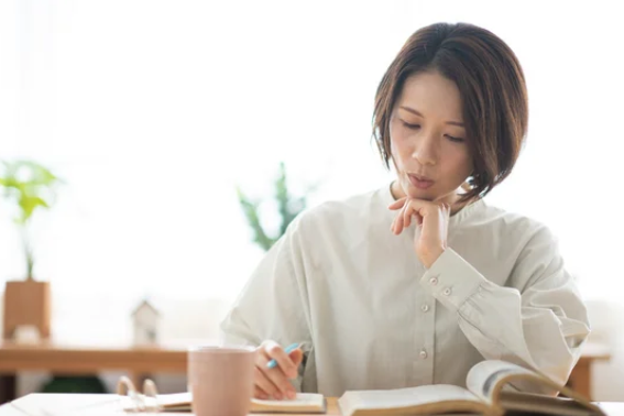 ノートと本を前に顎に手を添えて熟考する日本人女性 ─ Mani Padmeスピリチュアルの科学的根拠を探る学びの姿